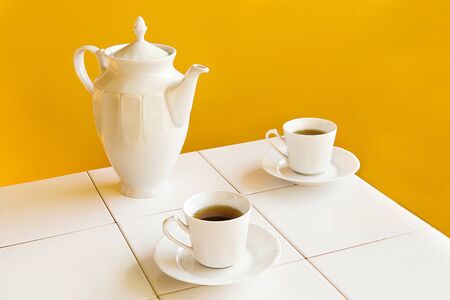 White tea cups and teapot on white tiles table over yellow background.の写真素材