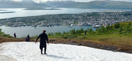 Snow on the top of the Mount Storsteinen in summer and panoramic view of Tromso city in Norway. Man slides down from snowy mountain in Tromso, Norway.の写真素材