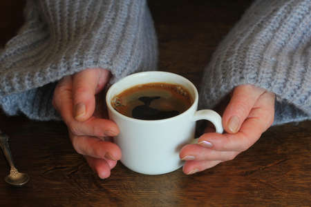 Woman's hands in sweater hold a cup of strong coffee on wooden table. Coffee fan top view background...