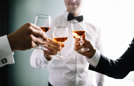 groom and his groomsmen friends in stylish suits drinking whiskey in hotel room, morning before the wedding preparation, emotional group of friends celebratingの写真素材