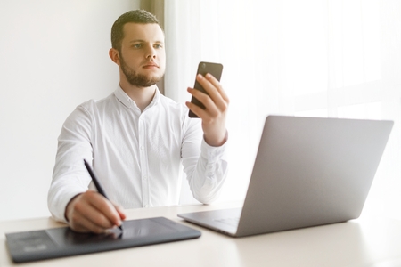 Young smiling bearded businessman is working on computer holding smartphone.の写真素材