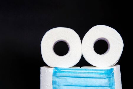 toilet paper rolls with a medical mask folded in the form of an emoticon, on a black background. concept of panic during coronavirus. copy spaceの写真素材