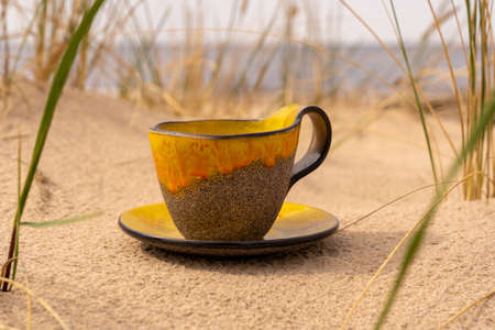 A clay cup with a saucer is placed in the sand of the beach between the bends.

A clay cup with a yellow glass glaze and a matching saucer is placed in the beach sand, by the sea.

Side view of the cup with a saucer.の写真素材