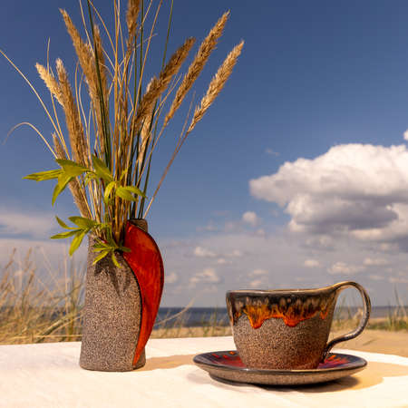 A clay ceramic vase and a cup with a saucer placed on a linen tablecloth on the sea beach.

The lower parts of the vase and mug are a layer of coarse sand and decorative reddish-orange glass glaze on the edges. Bent in a vaseの写真素材