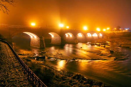 Autumn night view of an old brick bridge built in arches where fog rises from the riverold brick bridge over a wide river on a dark autumn night with fog and bridge lightingの写真素材