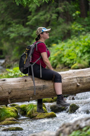 a break in a mountain hike, sitting on a log that has fallen over a stream, a woman looks around in summerの写真素材