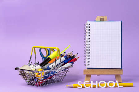 Metal shopping basket with school supplies on a lilac background. School shopping concept.