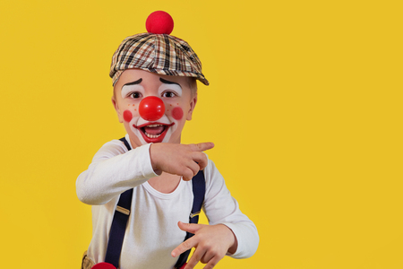 Child clown smile shows a finger copy space area on yellow background. Funny little clown with red nose. Concept birthday, day 1 april, party. Beautiful portrait kid jester in studio.の写真素材