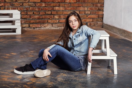 Brunette hipster teenager sitting floor. Stylish adolescent girl wears casual clothes, jeans jacket. Fashion pose, serious character. Behind young woman background brown brick wall. series photos.の写真素材