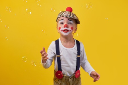 Funny kid clown looking up on bubbles, soft focus.Little boy clown in a costume, makeup,bright yellow background. Concept birthday,celebration,party.Young kid expression face calm, curious, sad.の写真素材