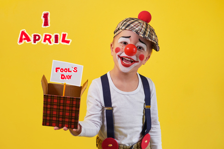 Beautiful portrait funny kid in celebration costume clown isolated on yellow background. Child clown smile keeping box. Concept birthday, day 1 april, party. Stylish emoticon jester kid 5 years.の写真素材