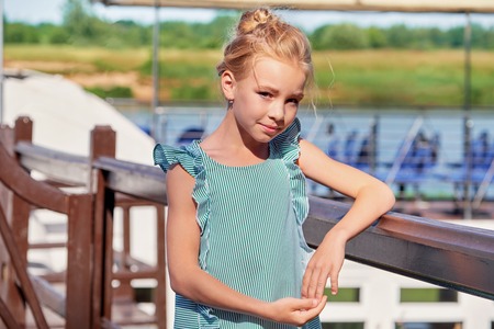 Portrait elegant young girl of 9-11 years outdoors, leaning her elbows on the railing. Cute child blonde smiling, sunny summer day resting on pier. Fashion kid concept. striped dress, marine style.の写真素材