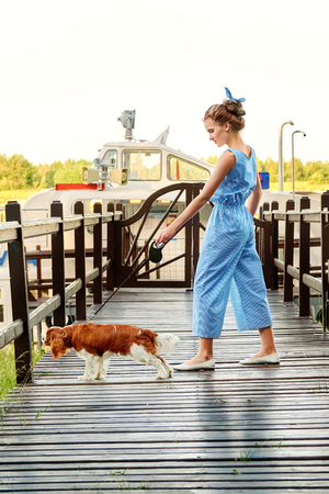 Fashion girl in striped blue overall posing with dog breed cocker spaniel. Beautiful young woman teenager standing on bridge pier. Designer collection.Fashion concept. Summer,day,outdoors.の写真素材