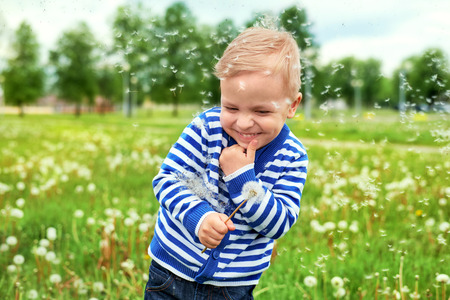 Happy smile kid standing in green grass dandelions. Emotion face caucasian child enjoyment summer. Portrait little boy in park outdoors. Joyful childhood. Dandelion seeds flying, green backgroundの写真素材
