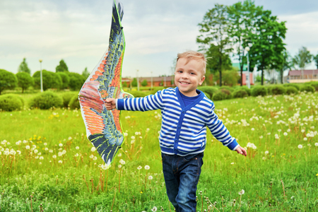 Happy little boy smiling running across park with kite bird flying. Child fun playing outdoors on summer or spring day.Joyful childhood. Lifestyle. Attractive kid actively recreation on nature, grass.の写真素材