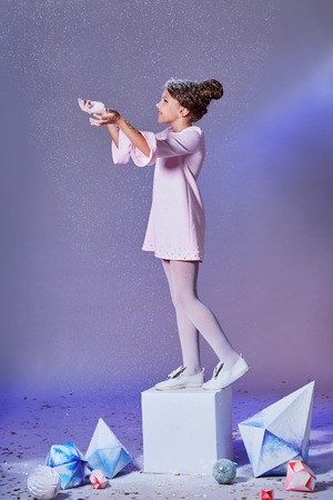 Beautiful young little child in elegance pink dress for party standing on a cube, catches snow by hand. Holidays, christmas, new year, x-mas, Fashion concept. Studio, purple background.の写真素材