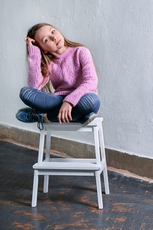 Stylish child girl sitting fashion pose on chair. Beautiful glamorous kid teenager model in pink sweater. casual,urban,street,youth style clothes. Studio shot, loft interior.の写真素材