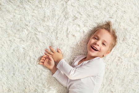 child funny home. Portrait happy little blond boy lying on soft bedspread bed, top view. Little kid laughing while looking at camera. copy space. top viewの写真素材