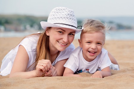 Mom and son smiling lie on sand of beach. Happy little boy son and mother summer. Portrait close-up young woman in hat together with cute blond kid having rest on outdoor, vacation.の写真素材