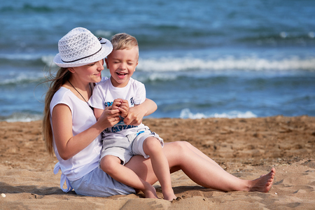 Young mother sitting on beach with son. Cute mom together child boy hugging, smiling, laugh, summer day. Happy childhood carefree playing on outdoor sand,sea, joy, fun. concept of holiday, vacation.の写真素材