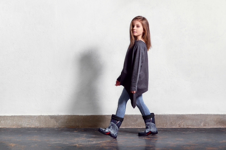 Fashion kid casual stylish clothes. Model child walks, pose. little girl poses near white wall, wears leggings, sweater, rubber boots. Full length portrait . Copy space.の写真素材