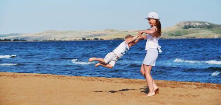 Young mother in summer clothes in hat together with son on beach. Carefree playing on outdoor sand,sea, joy, fun. Child boy, girl walking on coast. background panoramic,banner, copyspace.の写真素材