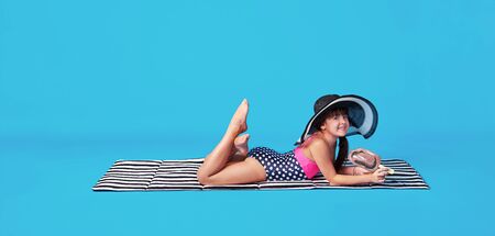 Attractive happy child lying in swimsuit, hat on isolated blue, colored background. Summer Vacation concept. Beautiful caucasian cheerful kid girl resting on towel beach.の写真素材