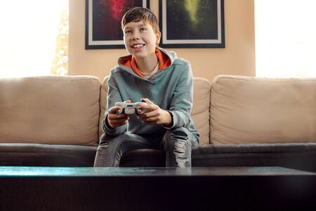 a cheerful boy sits on a sofa at home and plays on a game console, happy to win