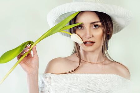portrait of a beautiful brunette girl in white clothes and a white hat with a tulip in hands on a white background, spring girlの写真素材