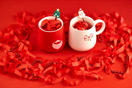 concept of holidays, christmas, winter and drinks - close-up of two cups of red and white color with christmas decorations on a red tableの写真素材