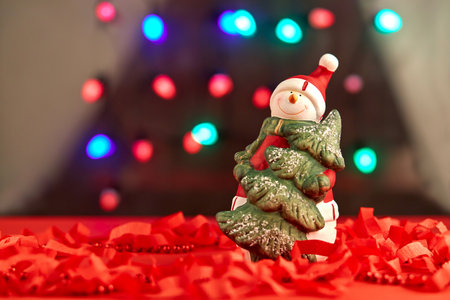 Stylish Christmas decoration, a cute ceramic souvenir-snowman with a Christmas tree stands on a New Years decorated window sill against the background of a window with blurry lightsの写真素材