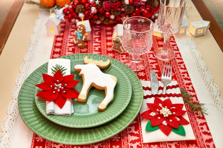 Christmas table setting with tree, garlands, poinsettia, wineglass, gingerbread cookies and lights on a wooden table covered with a red and white tableclothの写真素材