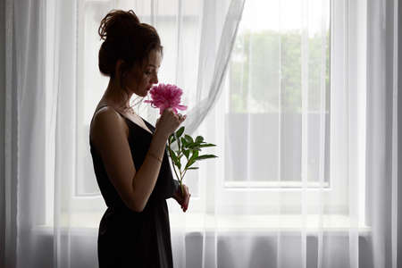 silhouette of a young sad woman in a dress with a peony in her hands on the background of a window, the concept of a lonely womanの写真素材