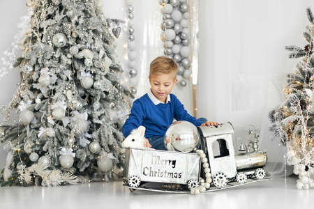 A small handsome blond boy sits near a Christmas tree in white tones and plays with a real white rabbit who sits on a toy train.の写真素材