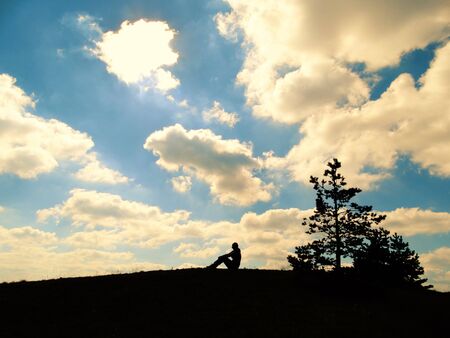 Silhouettes of a girl sitting over a mountain a lonely tree and a blue cloudy sky in the backgroundの写真素材