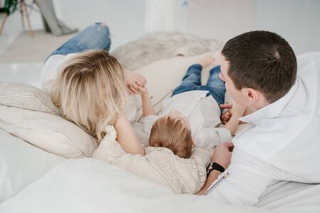 Mom, dad, son - lie on the bed in white shirts and jeans in a bright room - rear view from headの写真素材