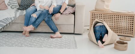 A child in blue jeans on the floor crawls into bag like a cat - in the background the legs of mom and dadの写真素材