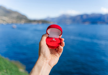 A hand holds a wedding ring made of white gold, platinum, silver, with a diamond in a red velvet box for a marriage proposal while traveling around Europe and the world - in Italy on Lake Comoの写真素材