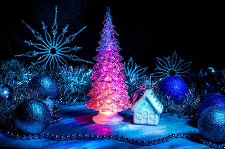Icy Christmas tree glowing with red light on a blue background. Surrounded with little house, blue balls, silver tinsel and snowflakes. Light brush. New Year and Christmas decoration.の写真素材