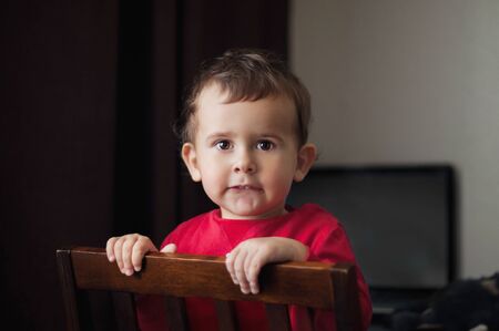 Cute 2 year old boy in a red shirt posing standing on a chair. Brown background.