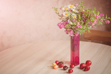 A bouquet of different rural flowers with camomiles in a pink vase on a table with ripe cherries. Summertime background.の写真素材