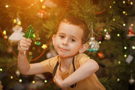 Little boy having fun near a christmas tree with a candy in shape of a fir tree at homeの写真素材