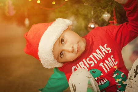 Little boy having fun under a christmas tree at home dressed in festive pajama and hood.