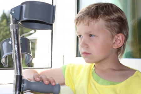 Seven years old boy with crutches sadly looks out of the windowの写真素材