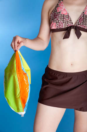 A woman in a two-piece swimsuit holds an uninflated (deflated) beach ball. Photographed with studio lights in front of a blue backdrop.の写真素材