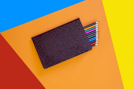 Closed notebook with a set of colored pencils on a colored background, top view