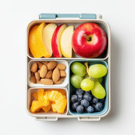 A top-down view of a neatly packed bento-style lunchbox filled with a variety of healthy snacks, including fresh apple slices, oranges, almonds, blueberries, and grapes, perfect for nutritious meals.の素材