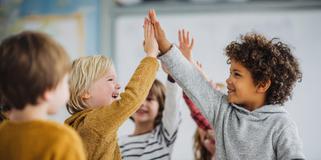 Two smiling students high-five each other in a vibrant classroom setting, with other children blurred in the background. This image perfectly captures themes of teamwork, achievement, friendship, and positive learning environments.の素材
