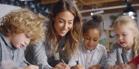 dedicated female teacher gently assists a group of children with their writing and drawing at a table in a bright, engaging classroom setting. This image perfectly illustrates interactive learning, personalized attention, and a supportive educational environment.の素材
