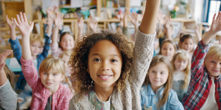 A happy young girl with curly hair confidently raises her hand and smiles at the camera, surrounded by a classroom full of other eager students also participating.の素材